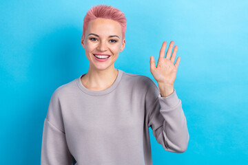 Photo of good mood pretty woman with dyed hairstyle dressed gray sweatshirt vawing arm to you say hi isolated on blue color background © deagreez