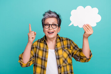 Photo portrait of lovely senior lady hold paper cloud finger point up wear trendy plaid yellow garment isolated on cyan color background