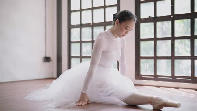 Young Asian ballet dancer in white tutu practicing ballet exercises bending forwards over her stretched body while sitting on the floor.