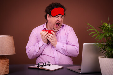 Funny fat man posing at a table with a laptop. Hard work online early morning Brown background.