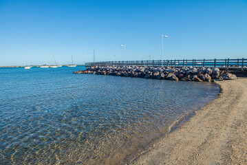 crystal clear water and a beach on the Malecon of La Paz Baja California Sur, in Mexico, with a pier in the background and a very calm sunny summer morning and clear blue sky, boats facing El Mogote.