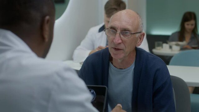 Elderly patient sits in clinic cafe with African American doctor. Professional medic discusses medical tests results with man. Digital tablet with MRI scan image. Hospital or medical center cafeteria.