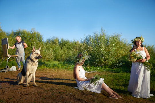 Adult female artist painting picture near water of river or lake, girls in white sundress and flower wreath posing in the holiday of Ivan Kupala and big dog shepherd in nature at sunset