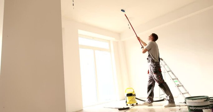 Man with a roller on long stick paints ceiling of house white. Repair of premises