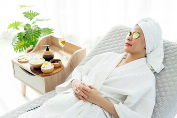 Relax young Asian woman in bathrobe, towel on a head, lying on the sofa bed with sliced cucumbers on her eyes and beauty treatment items for spa procedures such as essential oil and salt massage. - obrazy, fototapety, plakaty