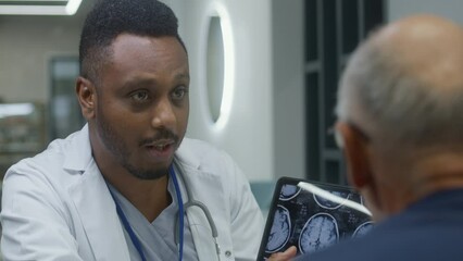 African American doctor sits in clinic cafe with elderly patient. Health care specialist talks to senior man, shows MRI or CT brain scan image using digital tablet. Hospital or medical center canteen.