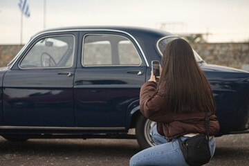 Una mujer con una campera marrón tomándole unas fotos a unos coches antiguos de colección con su celular - obrazy, fototapety, plakaty