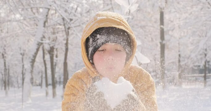 Little boy blows snow into the camera, joyful pink-cheeked child, sunny winter day, walking outdoors in cold winter, winter holidays