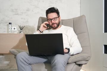 Young happy successful man in glasses working on laptop computer at home and talk on cell mobile phone