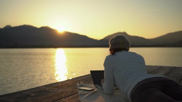 Freelance work choice concept: a young book author writes on her laptop computer while lying on the dock of a lake or sea at sunset, in a beautiful natural setting. Working remotely to live better