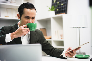 The manager of an Asian man sipping coffee on his desk in a radiant manner.
