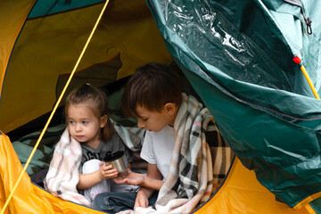Little boy and a little girl wrapped in plaid drinking hot tea sitting in a tent in the countryside. Concept of camping or refugeeism