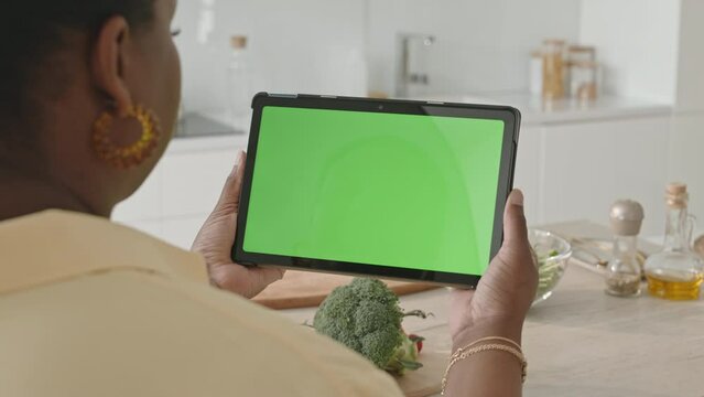 Over the shoulder shot of black woman holding digital tablet with chroma key green screen while preparing to cook meal at kitchen table at home
