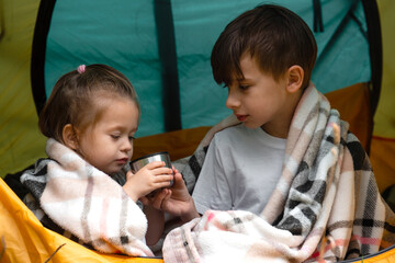 Little boy and a little girl wrapped in plaid drinking hot tea sitting in a tent in the countryside. Concept of camping or refugeeism
