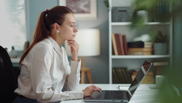 Woman is typing on a computer while sitting at her workplace desk, thoughtfully looks out the window and plans
