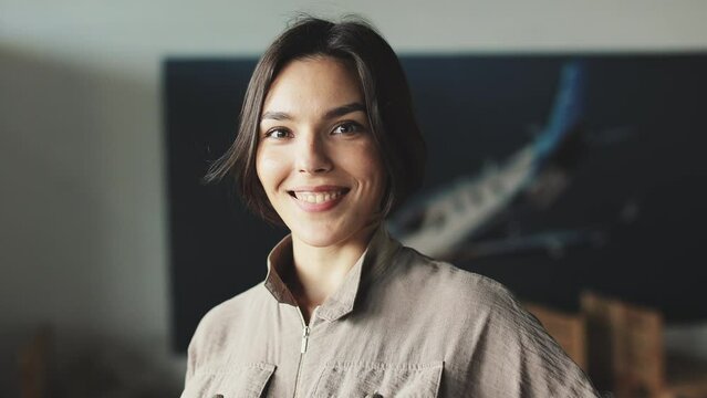 Close up of short-haired brunette posing against blurred background with large poster of aircraft at studio. Charismatic woman in coffee-colored outfit looking straight at camera and smiling.