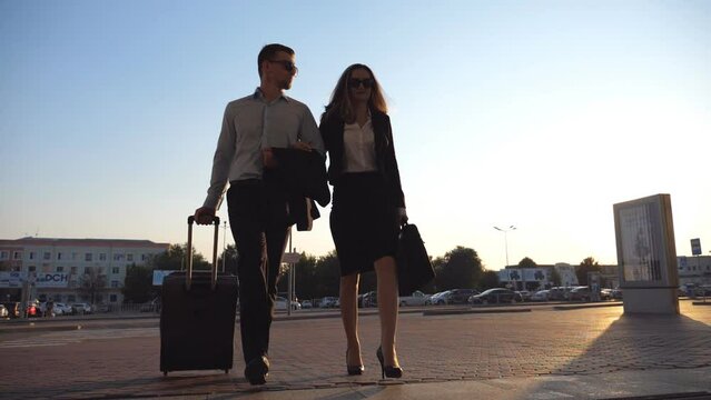 Businessman With His Female Colleague Walking Through Enter To Terminal And Roll Suitcase On Wheels. Business Man And Woman Stepping To The Airport Or Office Building Together. Slow Motion Close Up