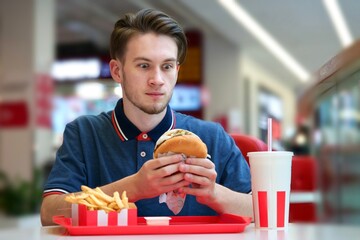 Young man eating fast food in the mall on food court. Burger, French fries and soda.