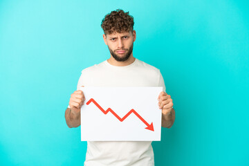 Young handsome caucasian man isolated on blue background holding a sign with a decreasing statistics arrow symbol with sad expression