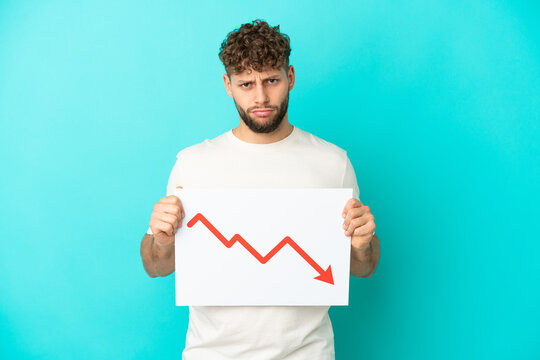 Young handsome caucasian man isolated on blue background holding a sign with a decreasing statistics arrow symbol with sad expression
