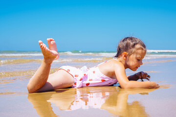Side view of tanned little girl child wearing white swimsuit, lying relaxing on beach, playing with...
