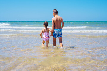 Back view of tanned preteen boy brother, little girl sister siblings standing walking in water on beach on sunny day.