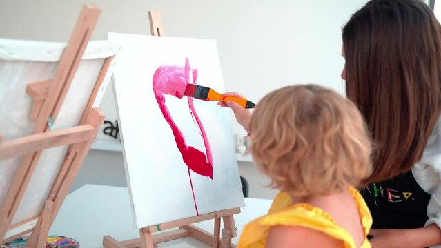 Teacher At Art School Helping Children in Art Class with painting. Woman teaches child to create and work with acrylic paint. Group of primary school kids learning how to draw and color help of their