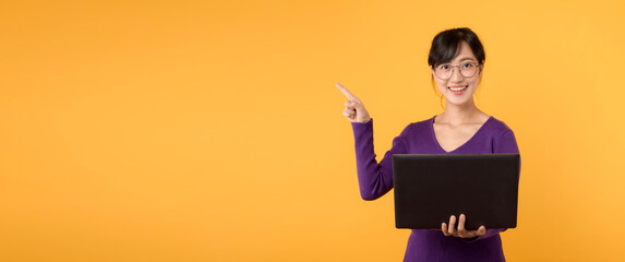 Asian female teacher 30s cute smile holding laptop pointing finger to free copy space, Wearing...