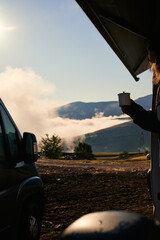 Side view of traveler in standing in camper van with hot drink and admiring picturesque scenery of mountain ridge in morning sunlight