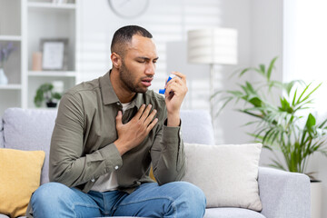 A young African American man is sitting on the couch at home and having an asthma attack. Holds his...