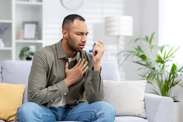 Foto op Canvas A young African American man is sitting on the couch at home and having an asthma attack. Holds his chest and breathes hard, blows an inhaler into his mouth © Tetiana