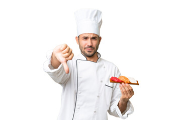 Young caucasian chef holding a sushi over isolated background showing thumb down with negative expression