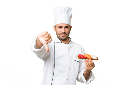 Young caucasian chef holding a sushi over isolated background showing thumb down with negative expression