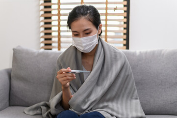 Young Asian woman wearing face mask have a cold and high fever while checking body temperature by...