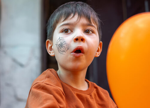 boy kid with orange balloons and spider web drawn on cheek playing outside, left building old door, wooden paint peeled door, child sitting on threshold.preschooler with sheet on head,like a ghost