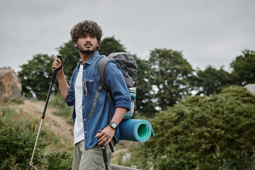 curly indian backpacker holding trekking poles on path in woods, hiker having adventure concept