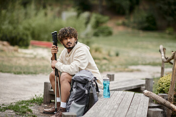 young indian tourist holding trekking poles near backpack while resting in forest