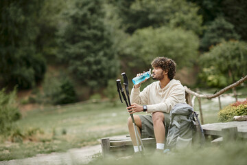 side view of indian tourist drinking water and holding trekking poles near backpack in forest