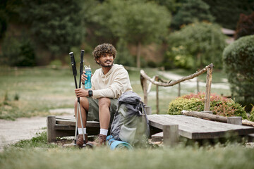 Fototapeta premium cheerful indian hiker holding trekking poles and sports bottle near backpack in forest