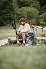 cheerful young indian hiker holding trekking poles and sports bottle near backpack in forest