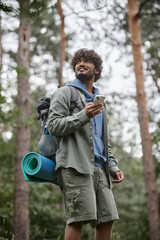 low angle view of cheerful young indian tourist with backpack using smartphone in forest