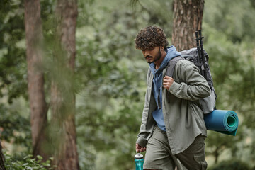 young indian tourist with backpack, trekking poles and sports bottle walking in forest