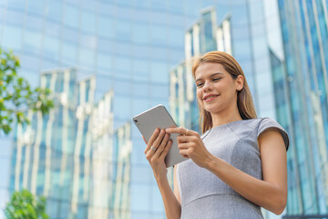 Fototapeta premium Business on the move. Digital banking on the go. Portrait of a young and successful white caucasian business woman using digital tablet standing in front of a modern cityscape of office buildings. 