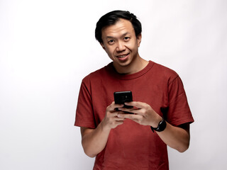 Fototapeta premium An Asian Indonesian man wearing a maroon-colored shirt is focused on replying to messages on his smartphone, isolated against a white background.