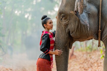 Young asian thai woman in traditional northeast costume pampering an elephant in a jungle. Thai lady posing with an elephant in a forest. - obrazy, fototapety, plakaty