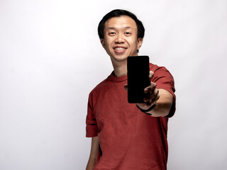 An Asian Indonesian man wearing a maroon-colored shirt is holding a smartphone and displaying it, isolated against a white background.