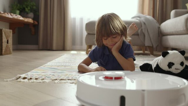 Portrait of cute smiling boy 5-7 years old with moving charming facial expressions lying on carpet watching robot vacuum cleaner. Technology makes life easier and comfortable. Autonomous room cleaning