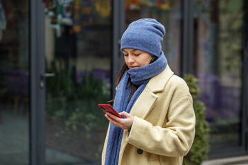 Woman in hat and scarf in beige coat sending text message using smartphone outside in city in winter