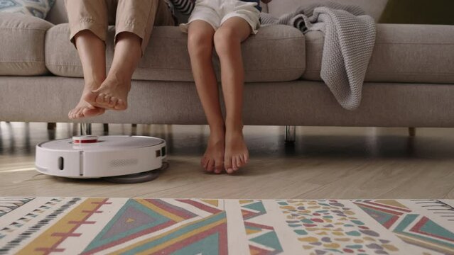 Vacuuming robot cleaning at home. Wireless robot vacuuming in the living room. Family mom and son raising their legs sitting on sofa while vacuum cleaner working. Domestic appliance and technology