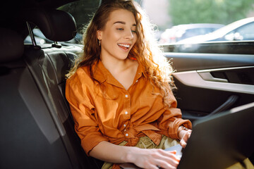 Young business woman with a modern laptop sitting in the back seat of a car. Successful startup, remote work. Concept of technology, freelancing.